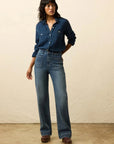 Woman wearing a blue denim shirt and jeans against a beige background