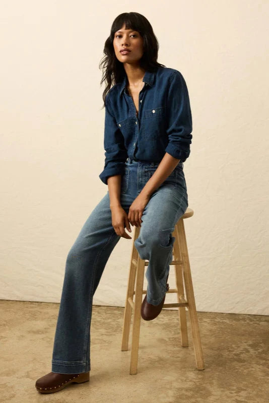 Woman sitting on a wooden stool wearing a blue shirt and jeans against a beige wall.