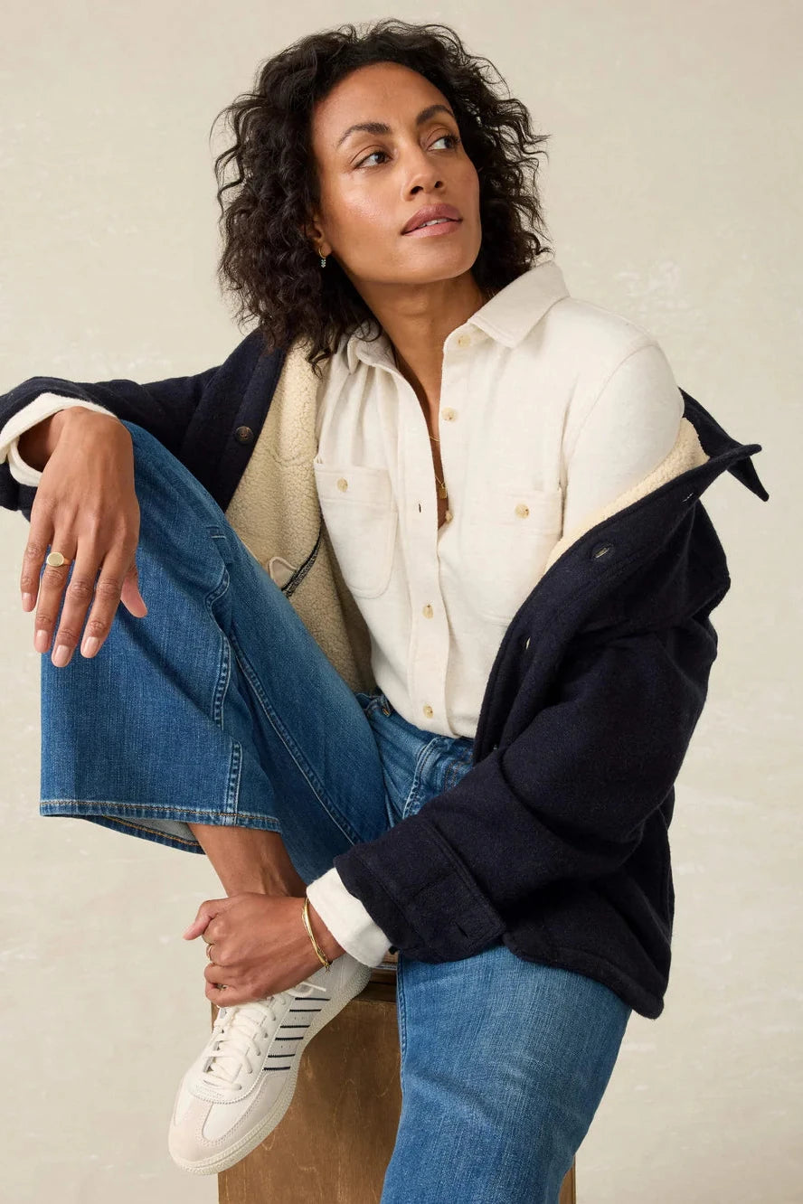 Woman wearing a navy coat, white shirt, and blue jeans against a beige background
