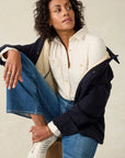 Woman wearing a navy coat, white shirt, and blue jeans against a beige background