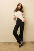 Woman wearing a white sweater with gray sleeves and black pants against a beige wall.