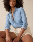 Woman wearing a light blue denim shirt and white shorts sitting on a chair.