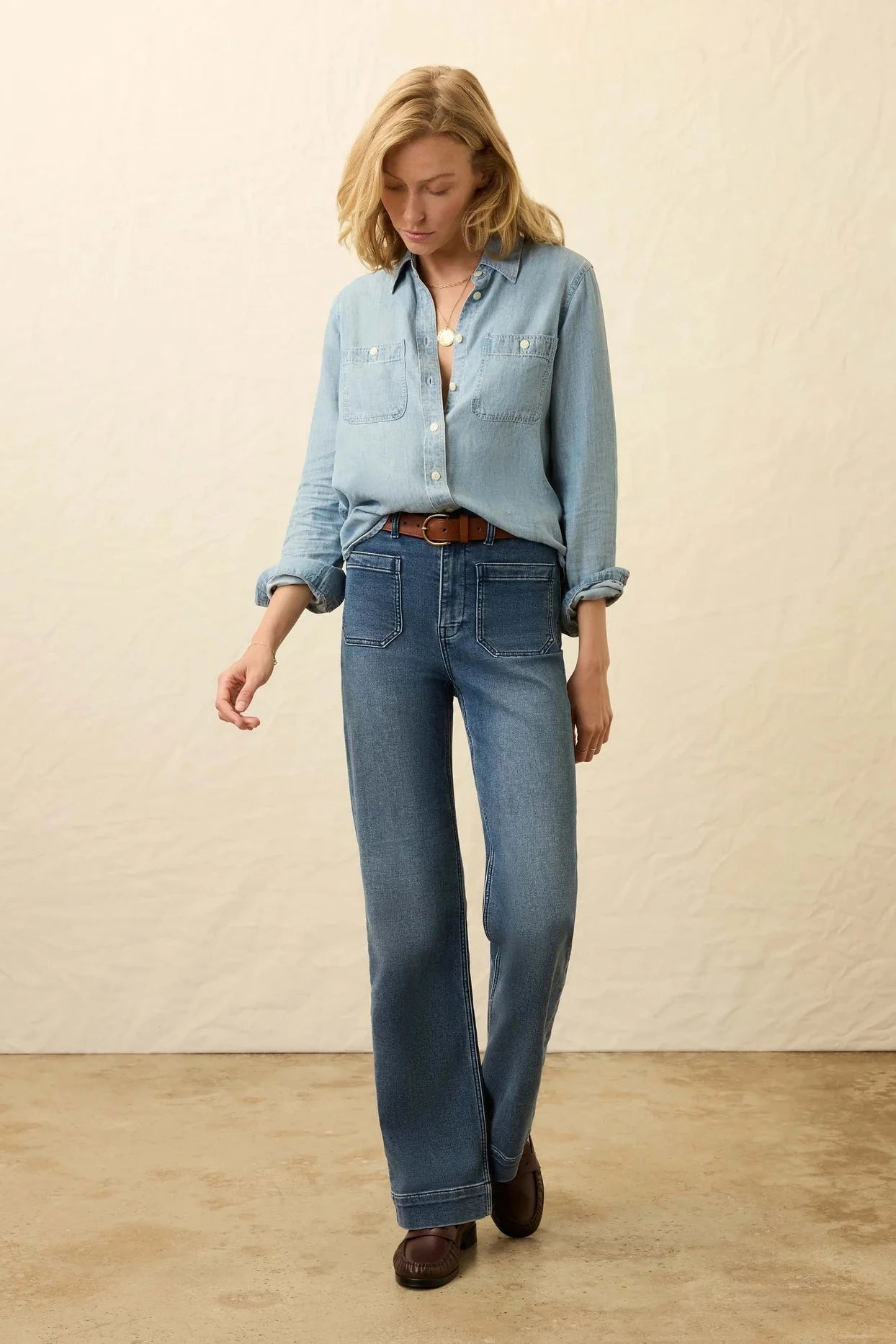 Woman wearing a light blue denim shirt and jeans against a beige background