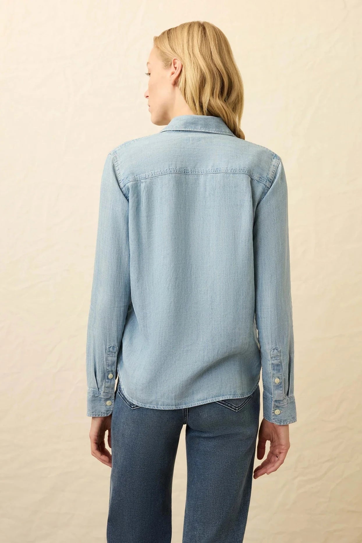 Person wearing a light blue denim shirt and jeans on a beige background