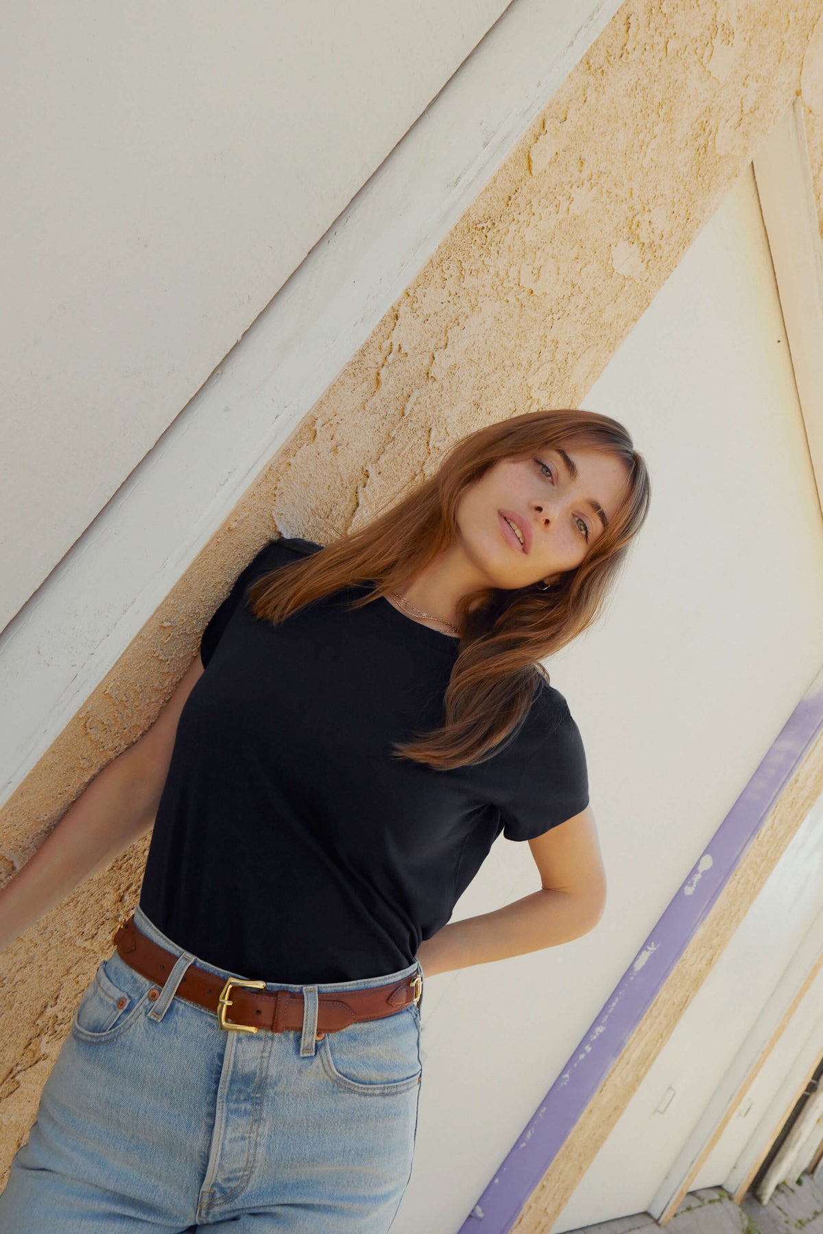 Woman wearing a black t-shirt and blue jeans standing against a textured wall.