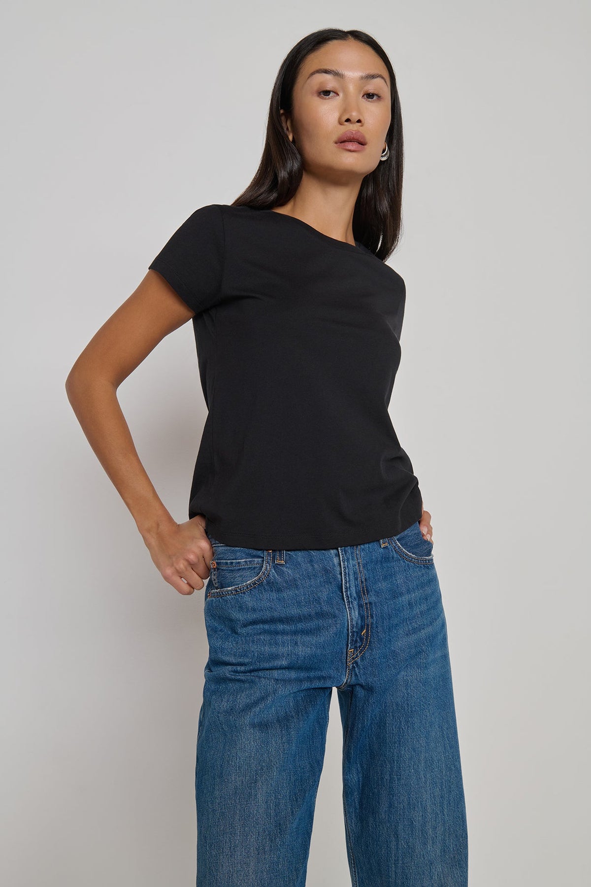 A woman modeling a black short-sleeve crewneck t-shirt paired with blue jeans, standing against a plain light gray studio background.