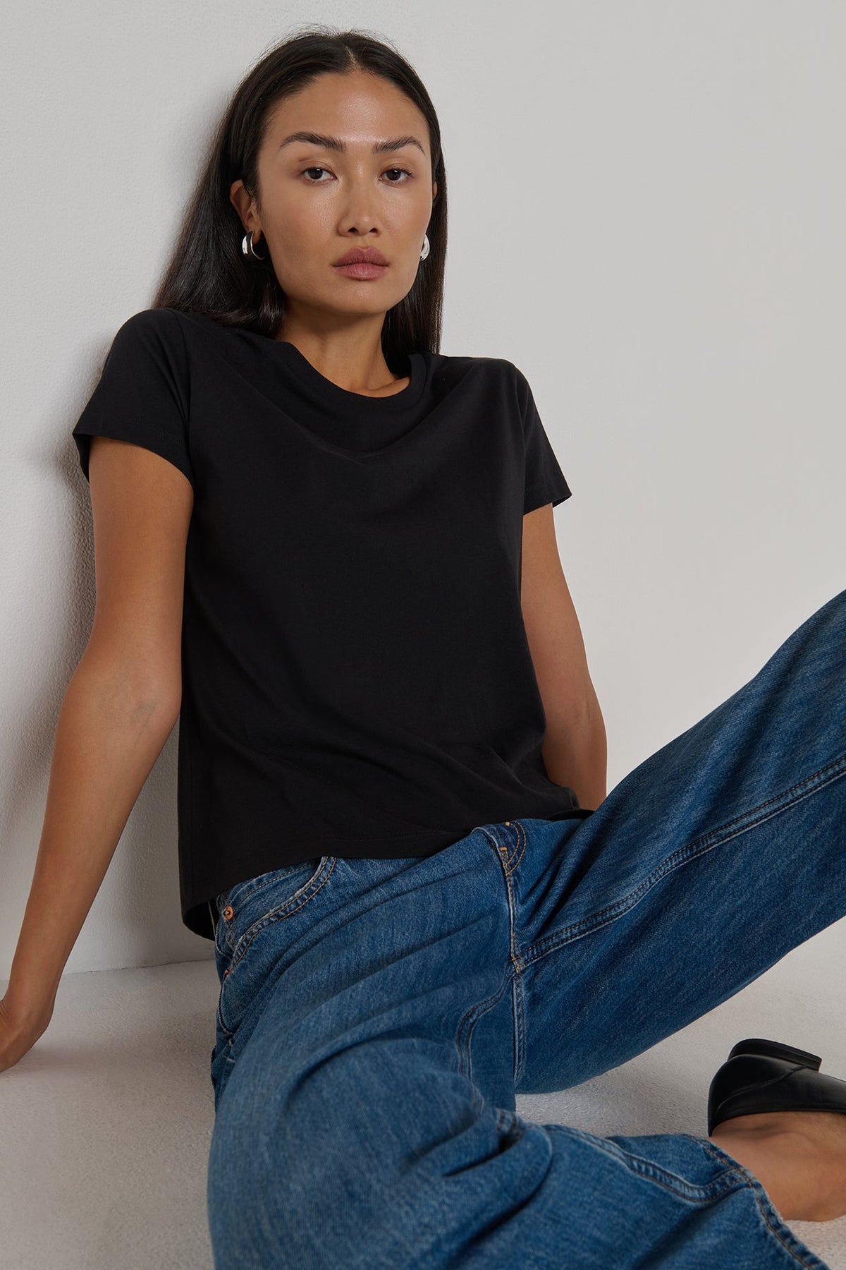 Woman wearing a black t-shirt and blue jeans sitting against a plain background