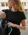 A woman wearing a black short-sleeve crewneck t-shirt and blue jeans carries a black leather shoulder bag and a green notebook in an outdoor urban setting.