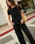 A woman wearing a black short-sleeve t-shirt and black wide-leg pants, carrying a tan woven shoulder bag, walking on a street crosswalk.