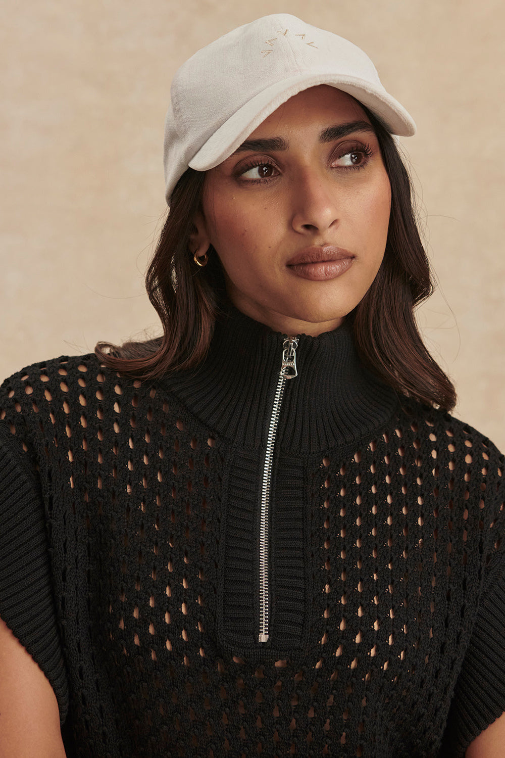 Woman wearing a black sleeveless sweater with a zipper and a beige cap against a beige background