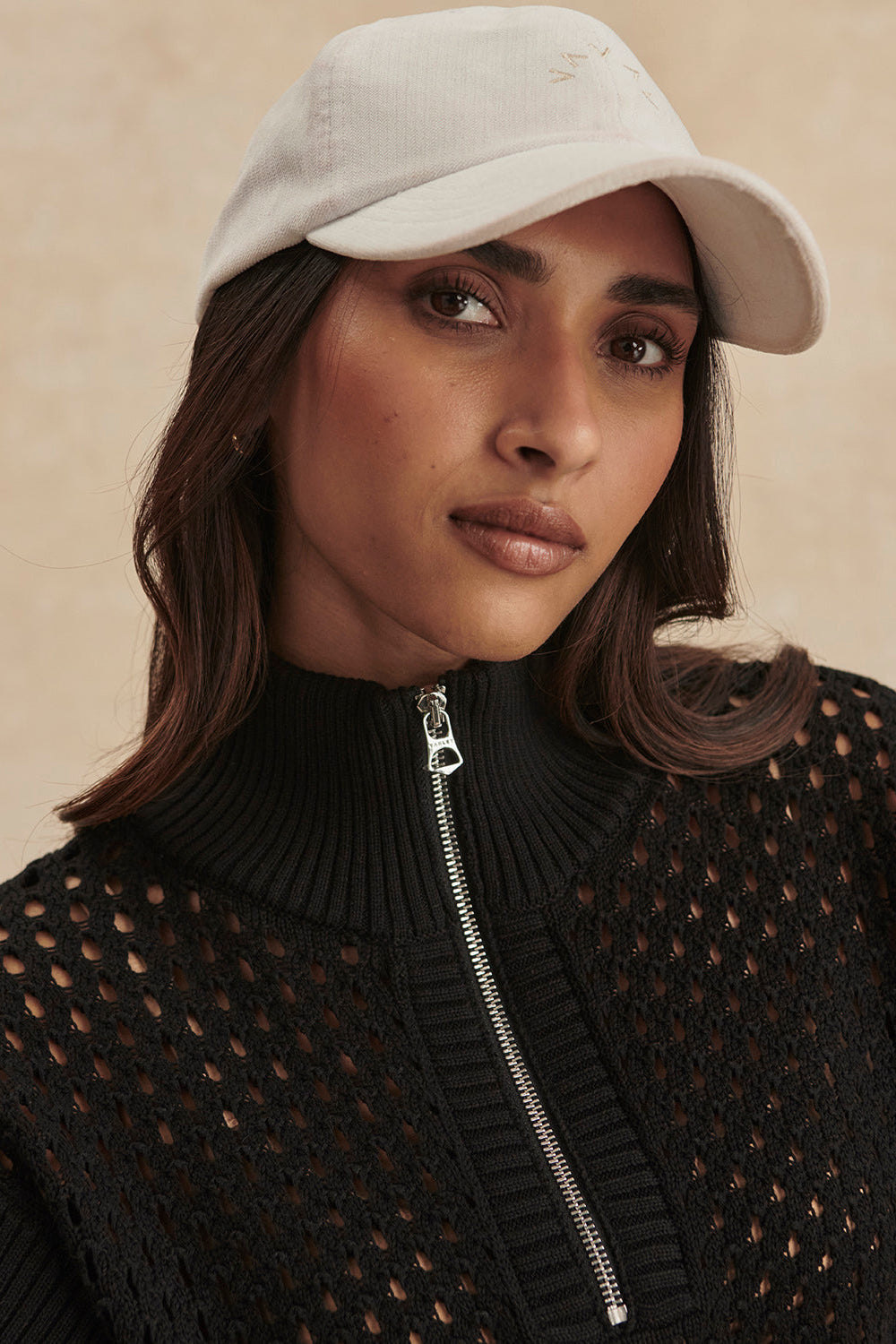 Woman wearing a beige cap and black textured sweater with a zipper.