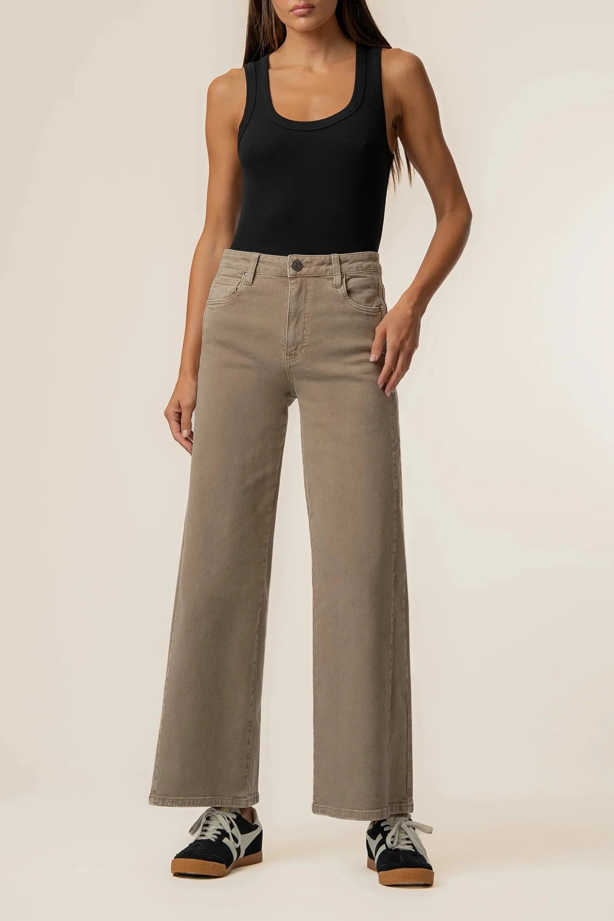 A model wearing a pair of high-waisted, wide-leg taupe jeans, paired with a black tank top and black sneakers, standing against a plain off-white studio background.