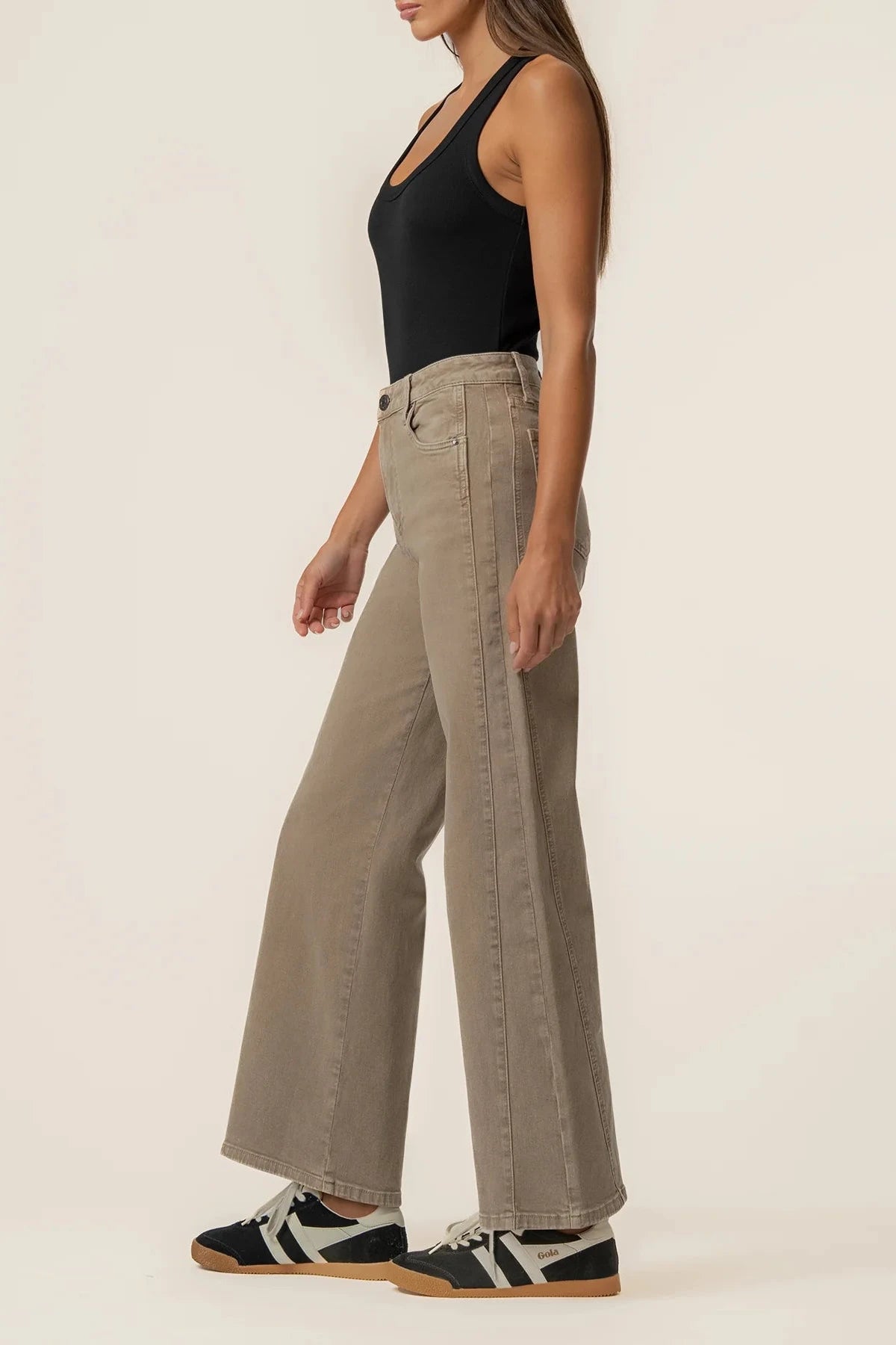 A woman shown from the side wearing high-waisted, wide-leg taupe pants, a black tank top, and black-and-white sneakers against a plain off-white studio background.