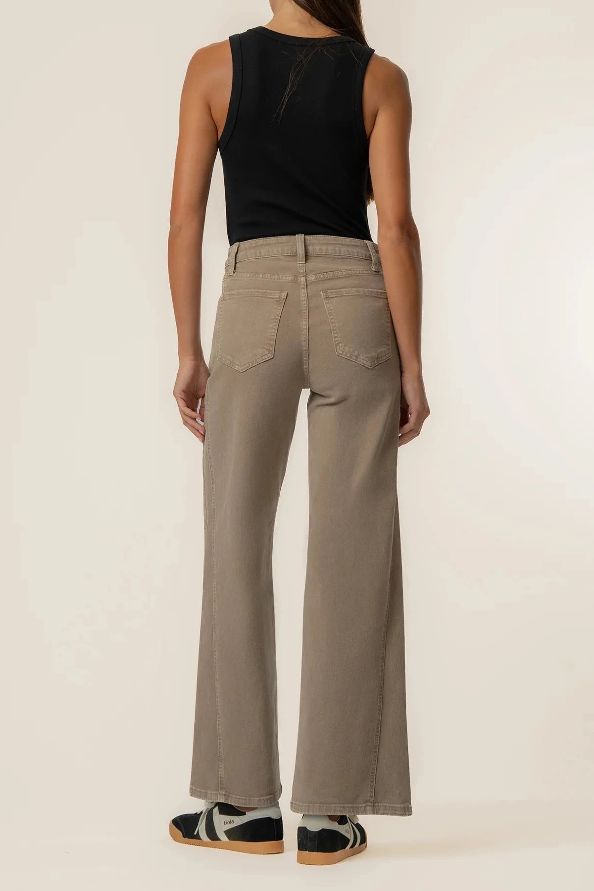 A model shown from the back wearing high-waisted, wide-leg taupe pants, a black sleeveless top, and black-and-white sneakers against a plain off-white studio background.