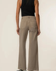 A model shown from the back wearing high-waisted, wide-leg taupe pants, a black sleeveless top, and black-and-white sneakers against a plain off-white studio background.