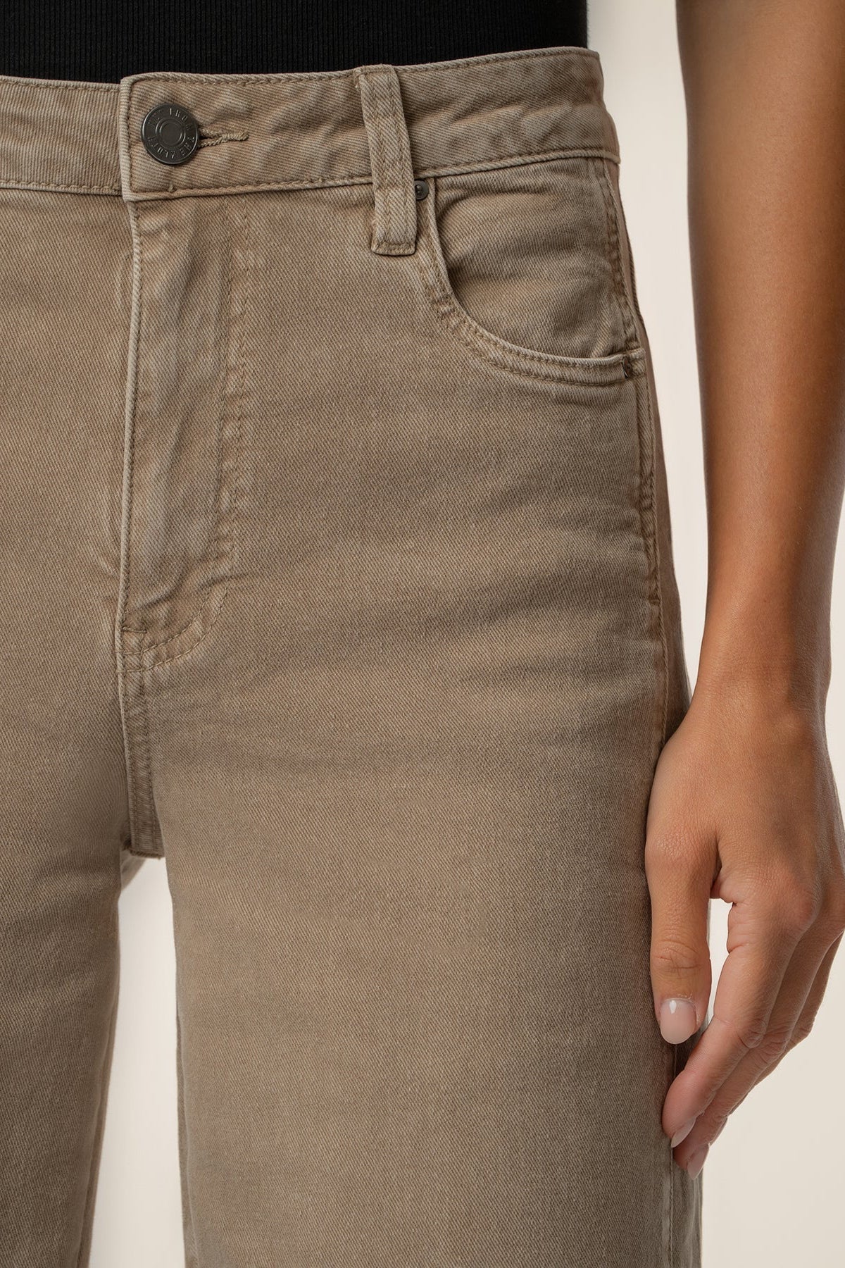 Close-up of a person wearing light brown or taupe denim jeans, showing the front waistband, button, belt loops, right pocket, and hand resting near the thigh.