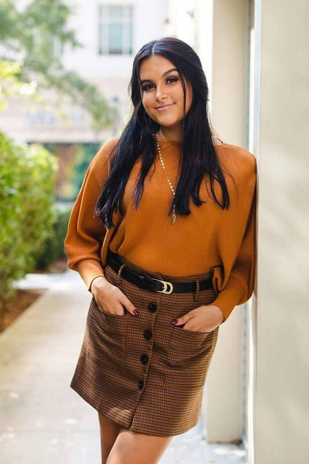 Woman in a mustard yellow top and brown skirt standing outdoors.