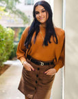 Woman in a mustard yellow top and brown skirt standing outdoors.