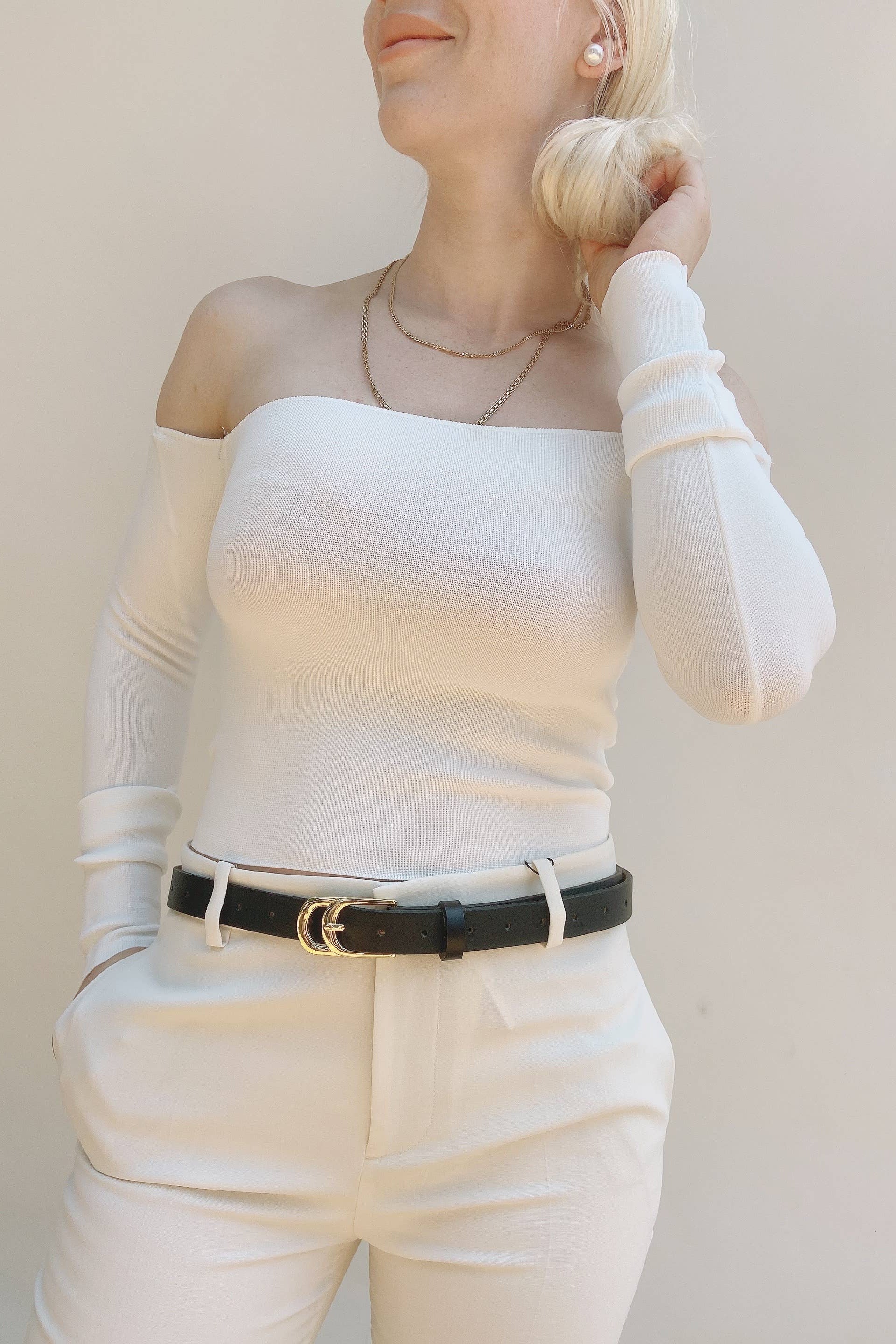Person wearing a white long-sleeve top and beige pants with a black belt against a plain background