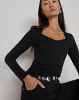 A woman modeling a black long-sleeve scoop-neck top with black jeans and a silver belt, posing against a light gray studio background.