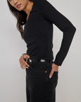 A woman modeling a fitted black long-sleeve top with ruching, paired with black jeans and a black belt, against a plain light gray studio background.