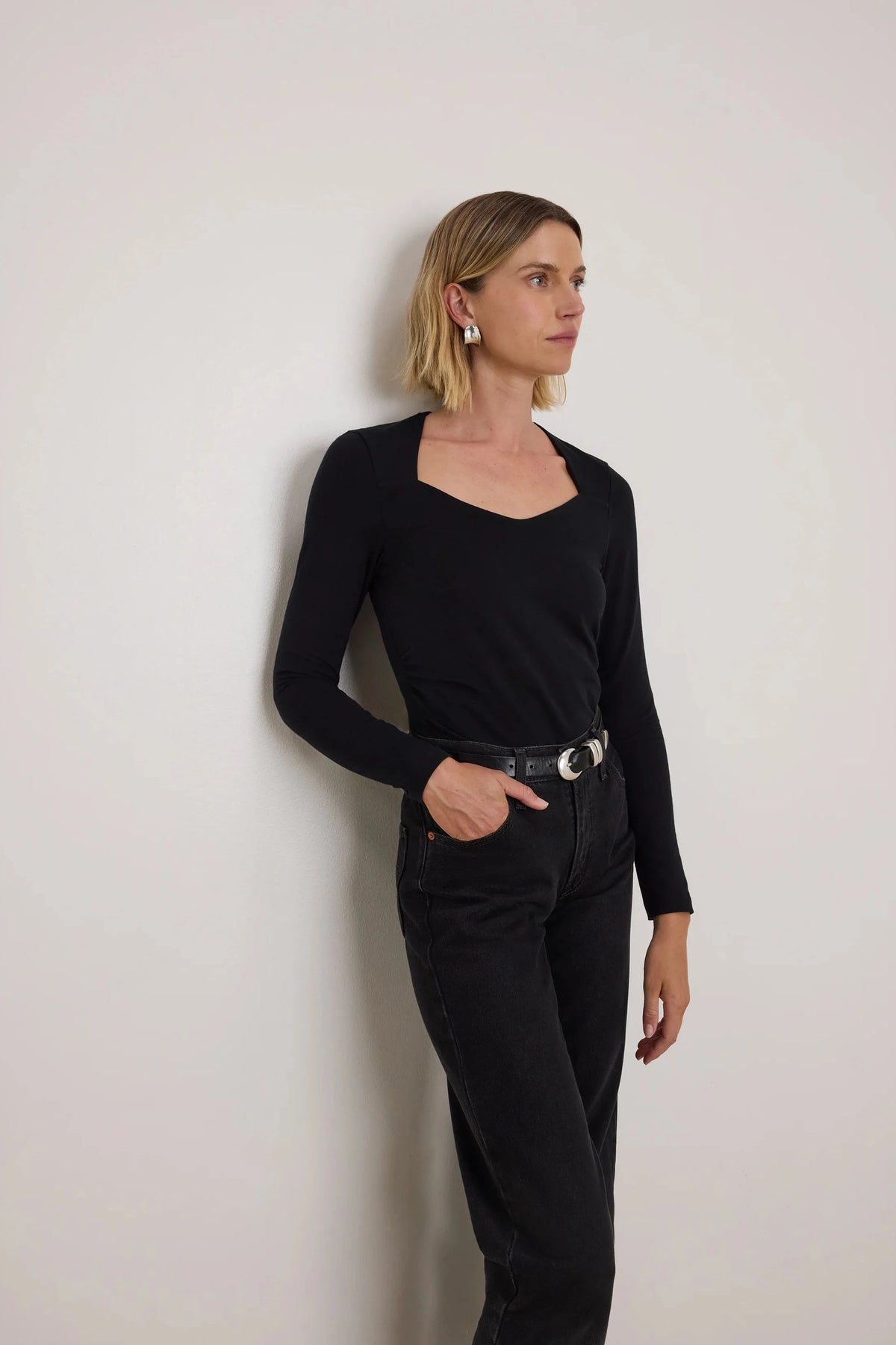 A woman with blonde hair stands against a plain light gray wall, wearing a black long-sleeve scoop-neck top tucked into black jeans with a black belt featuring silver hardware.
