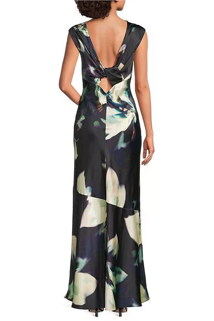 A sleeveless floor-length dress shown from the back on a model, featuring a deep V-neckline with a large bow detail and a dark abstract floral print in black, green, and cream.
