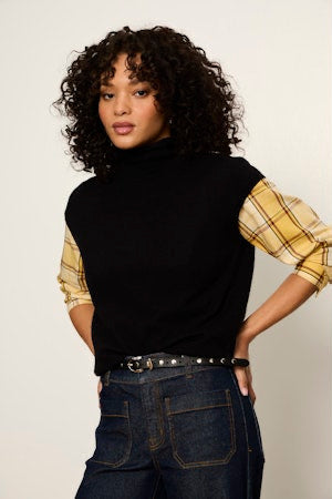 A model with curly dark hair wearing a black sleeveless mock-neck top with yellow plaid shirt sleeves, tucked into dark blue jeans with a studded belt.