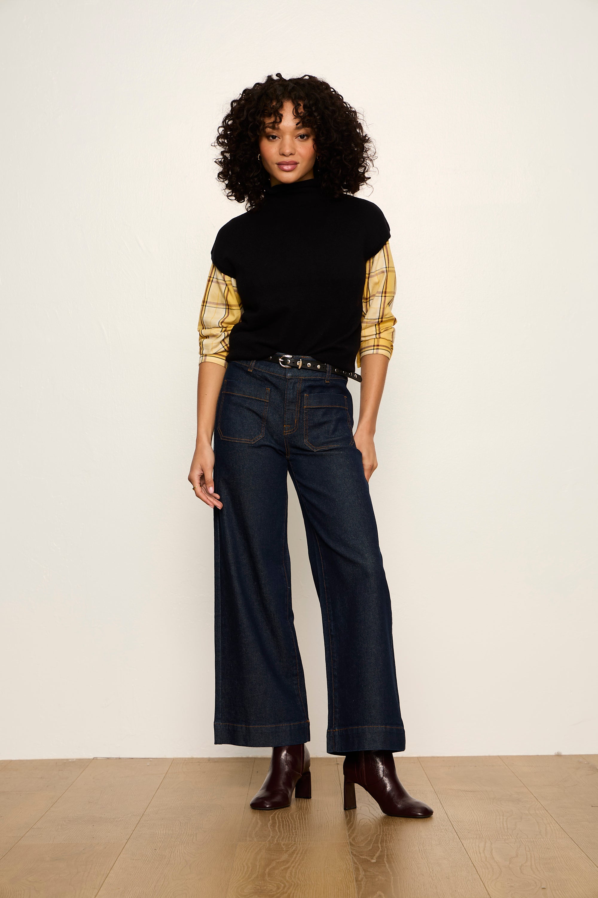 Woman wearing a black turtleneck, yellow long-sleeve shirt, and dark jeans against a white background