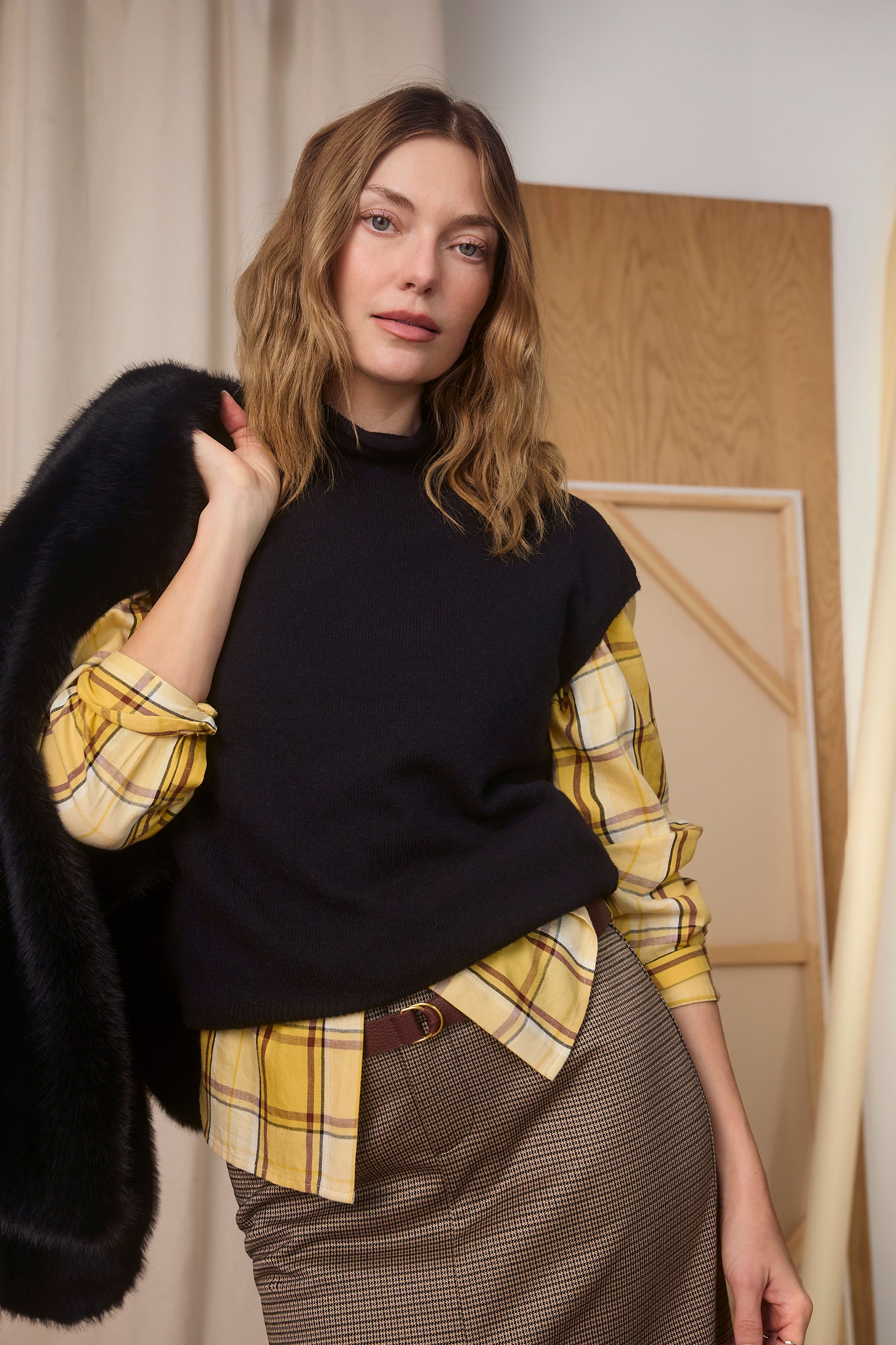 Woman wearing a black sweater over a yellow plaid shirt with a black coat, standing indoors.