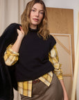 Woman wearing a black sweater over a yellow plaid shirt with a black coat, standing indoors.