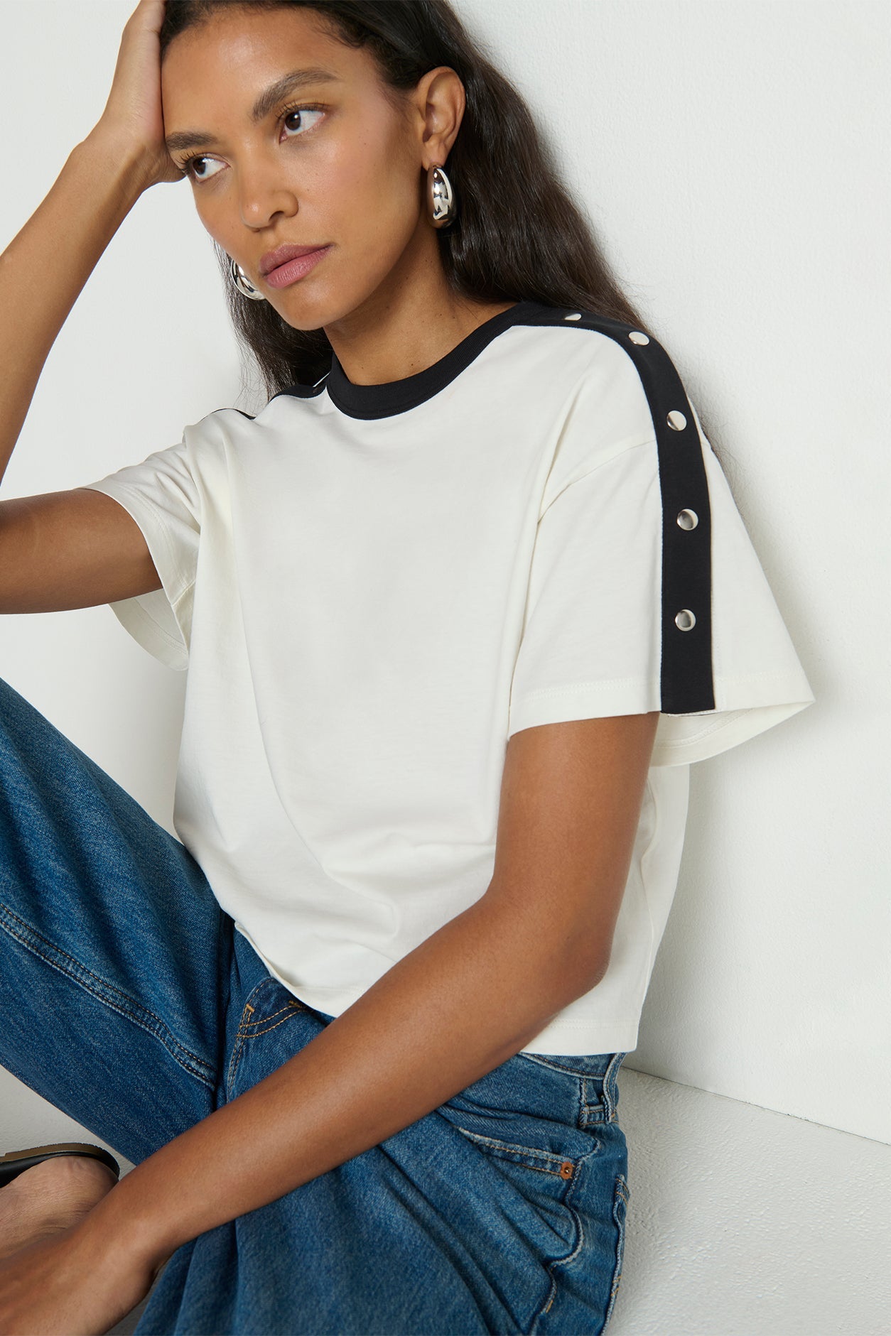 Woman wearing a white t-shirt with black trim and buttons, sitting against a white background