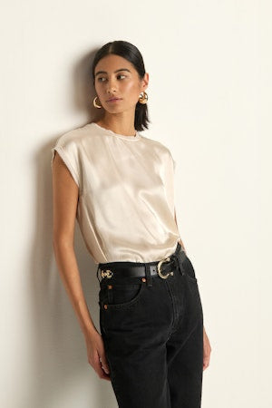 Woman wearing a beige blouse and black pants against a white wall