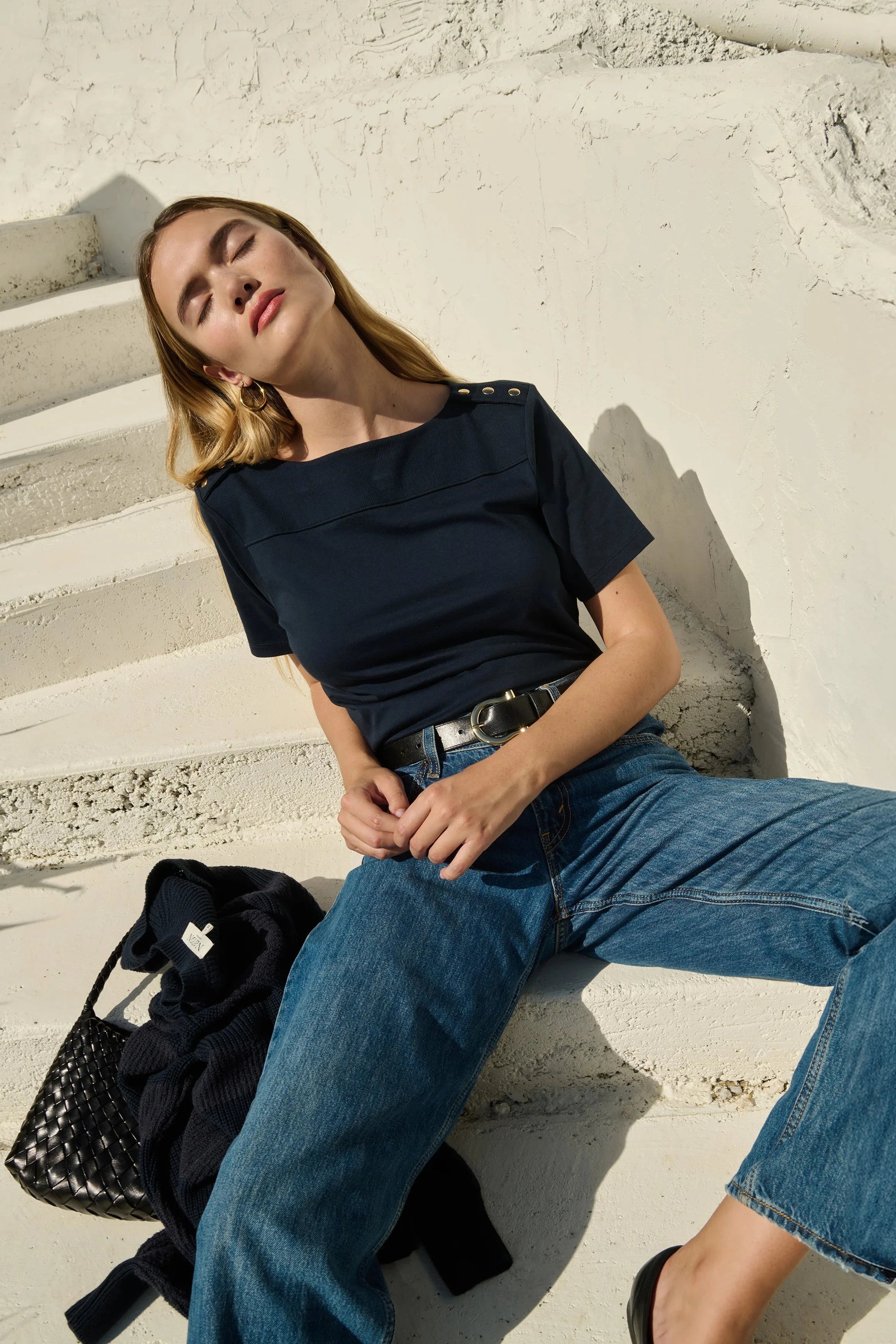 Woman sitting on steps wearing a black top and blue jeans with a black bag beside her.