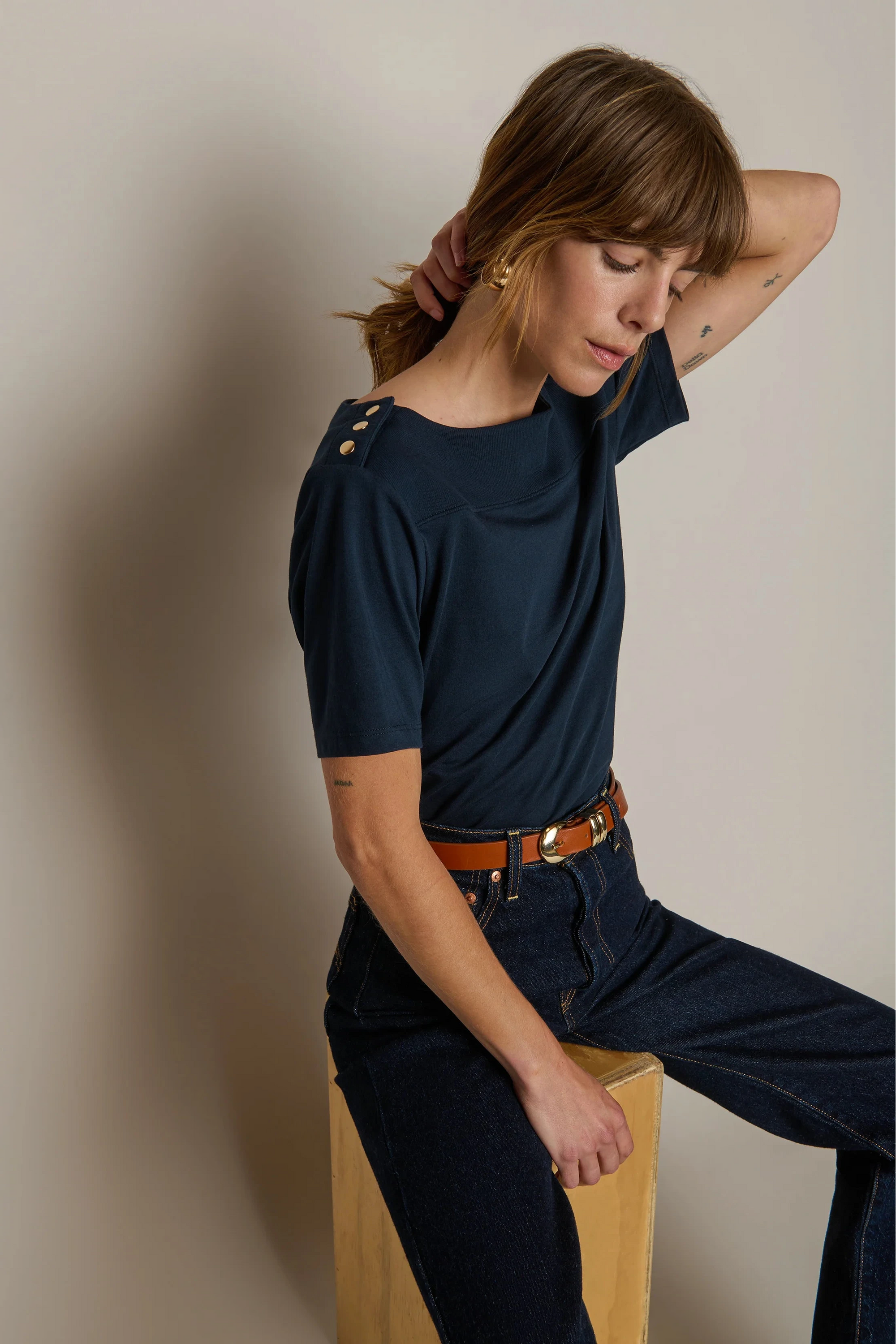 Woman wearing a navy blue top and jeans sitting on a wooden block against a beige wall.