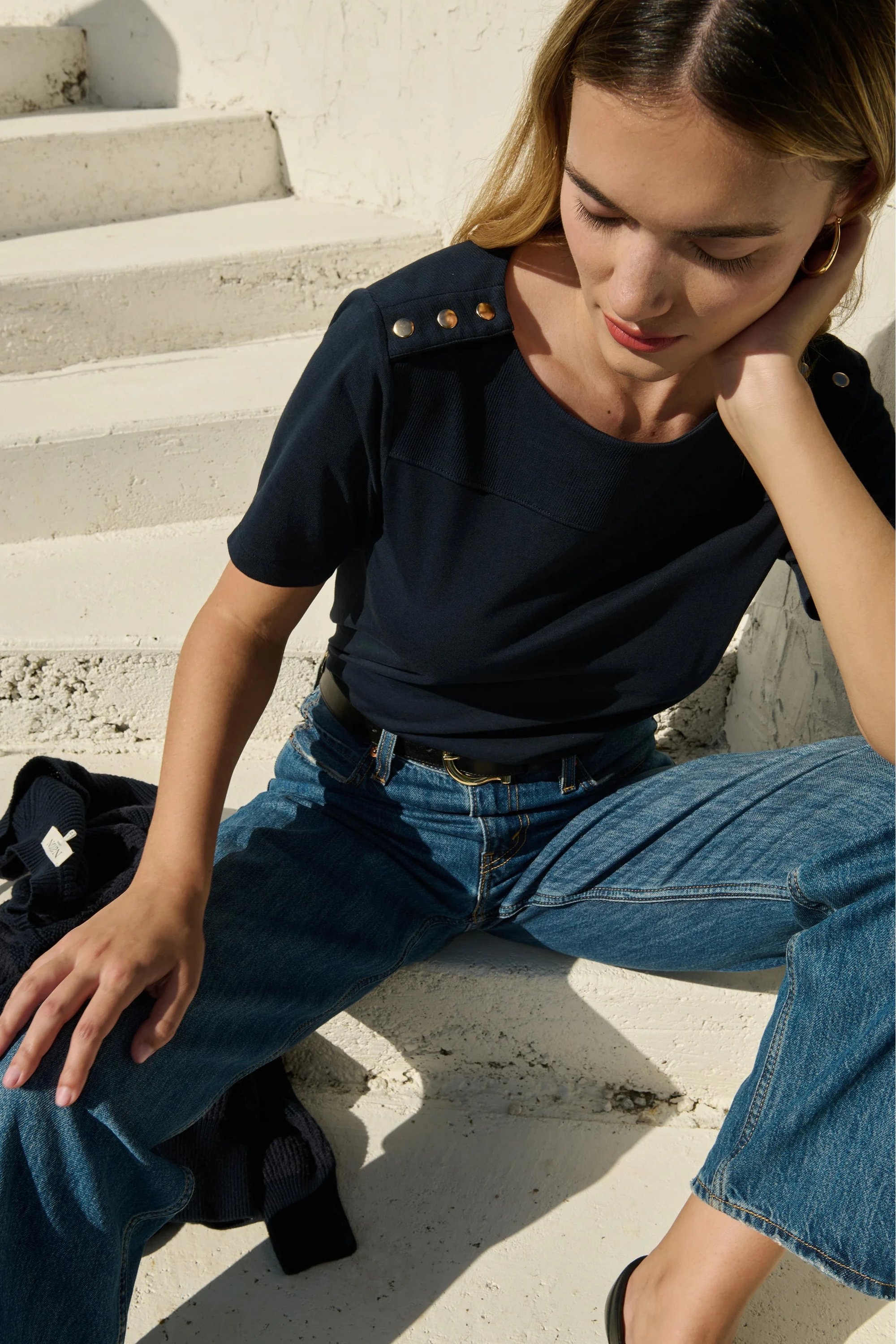 Woman sitting on steps wearing a black top and blue jeans with a black bag beside her.