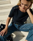 Woman sitting on steps wearing a black top and blue jeans with a black bag beside her.
