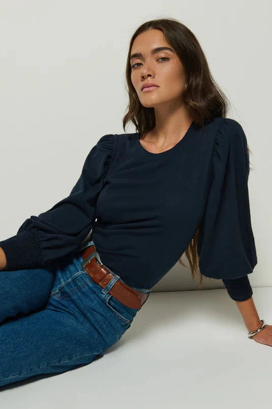 Woman wearing a navy blue top and jeans sitting on a white floor against a white wall.