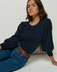 Woman wearing a navy blue top and jeans sitting on a white floor against a white wall.