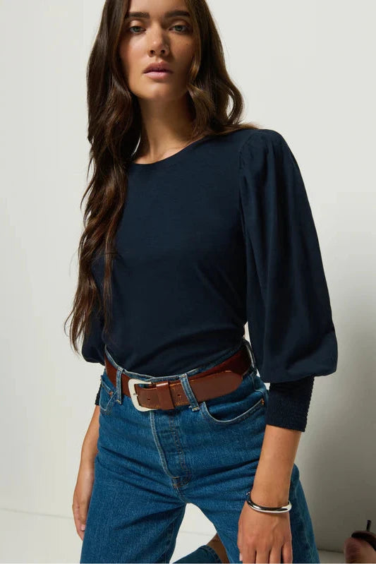 Woman wearing a navy blue top and blue jeans with a brown belt against a plain background