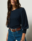 Woman wearing a navy blue top and blue jeans with a brown belt against a plain background