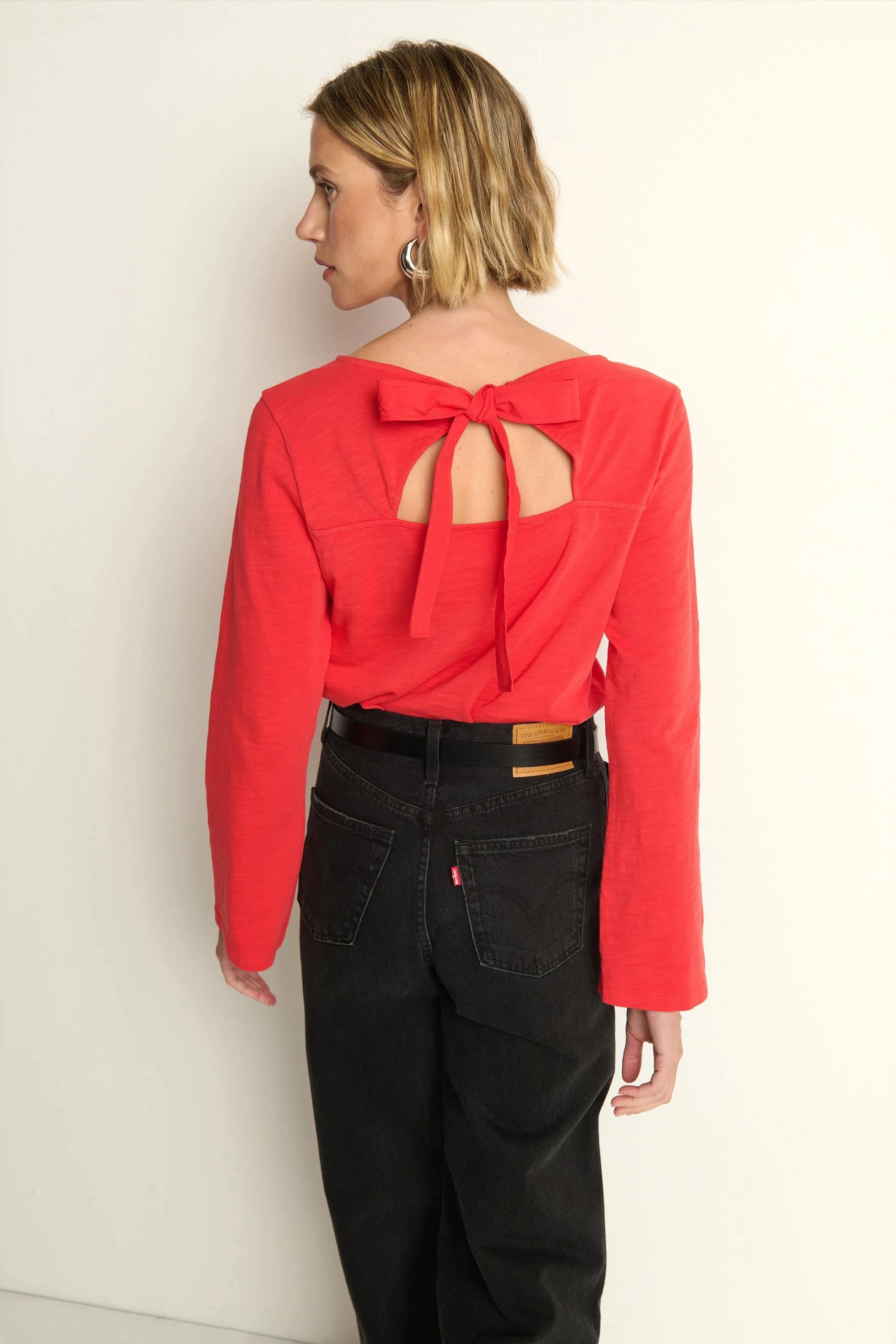 Woman wearing a red blouse with a bow detail on the back against a white wall