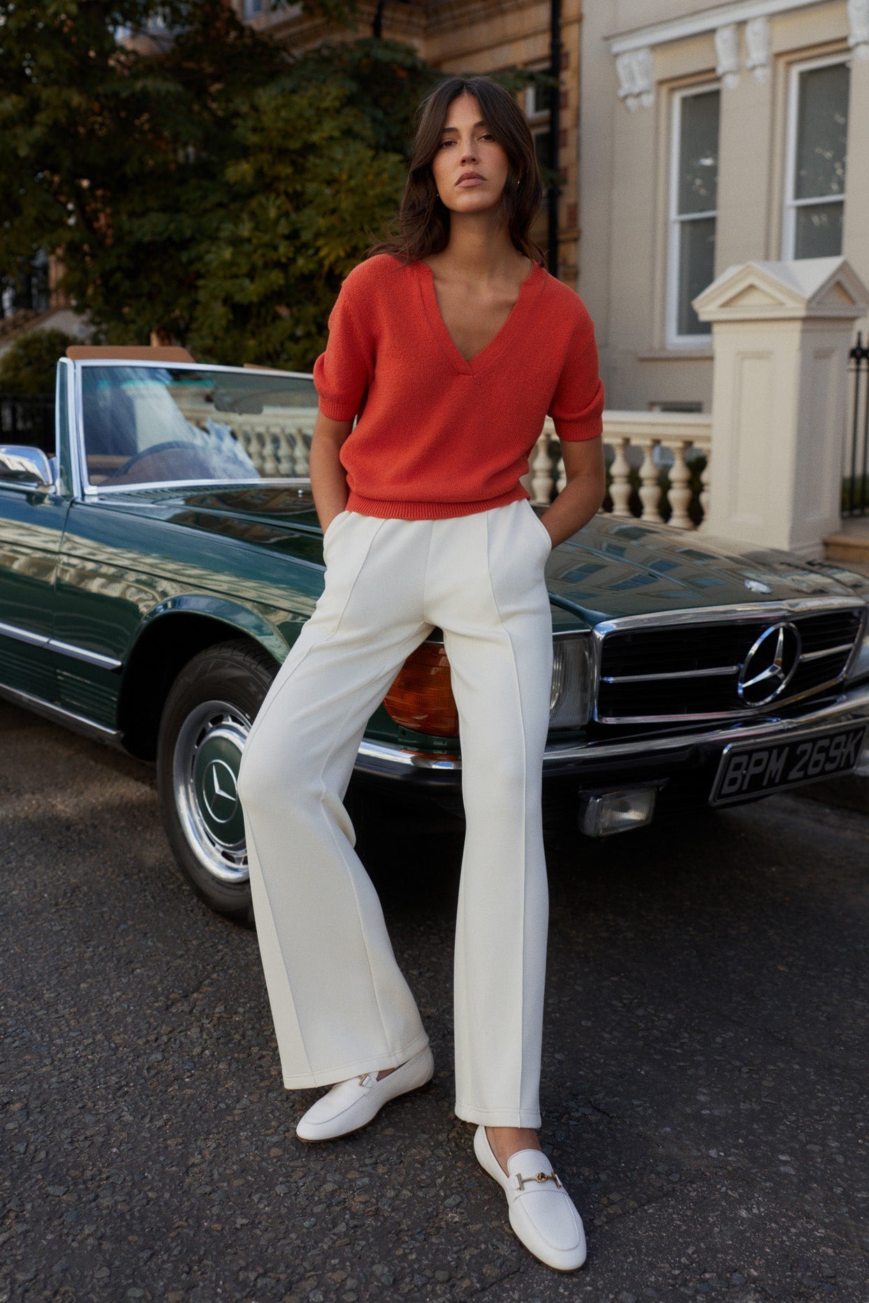 Woman in red sweater and white pants standing in front of a vintage car.