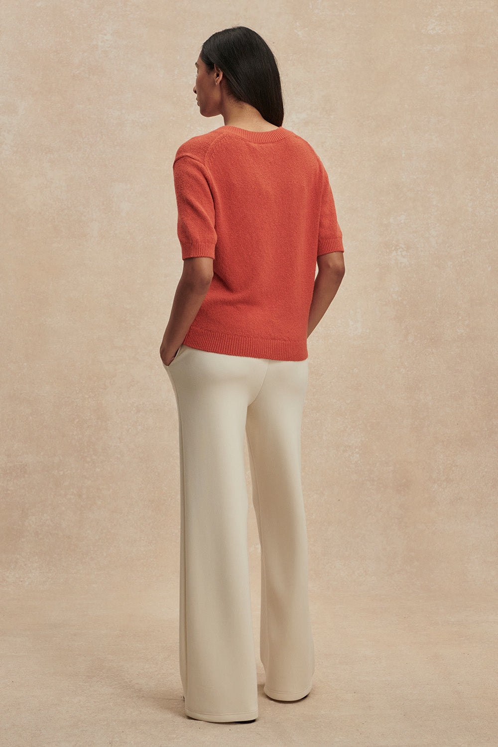 Person wearing an orange sweater and white pants against a beige background
