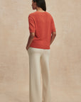 Person wearing an orange sweater and white pants against a beige background