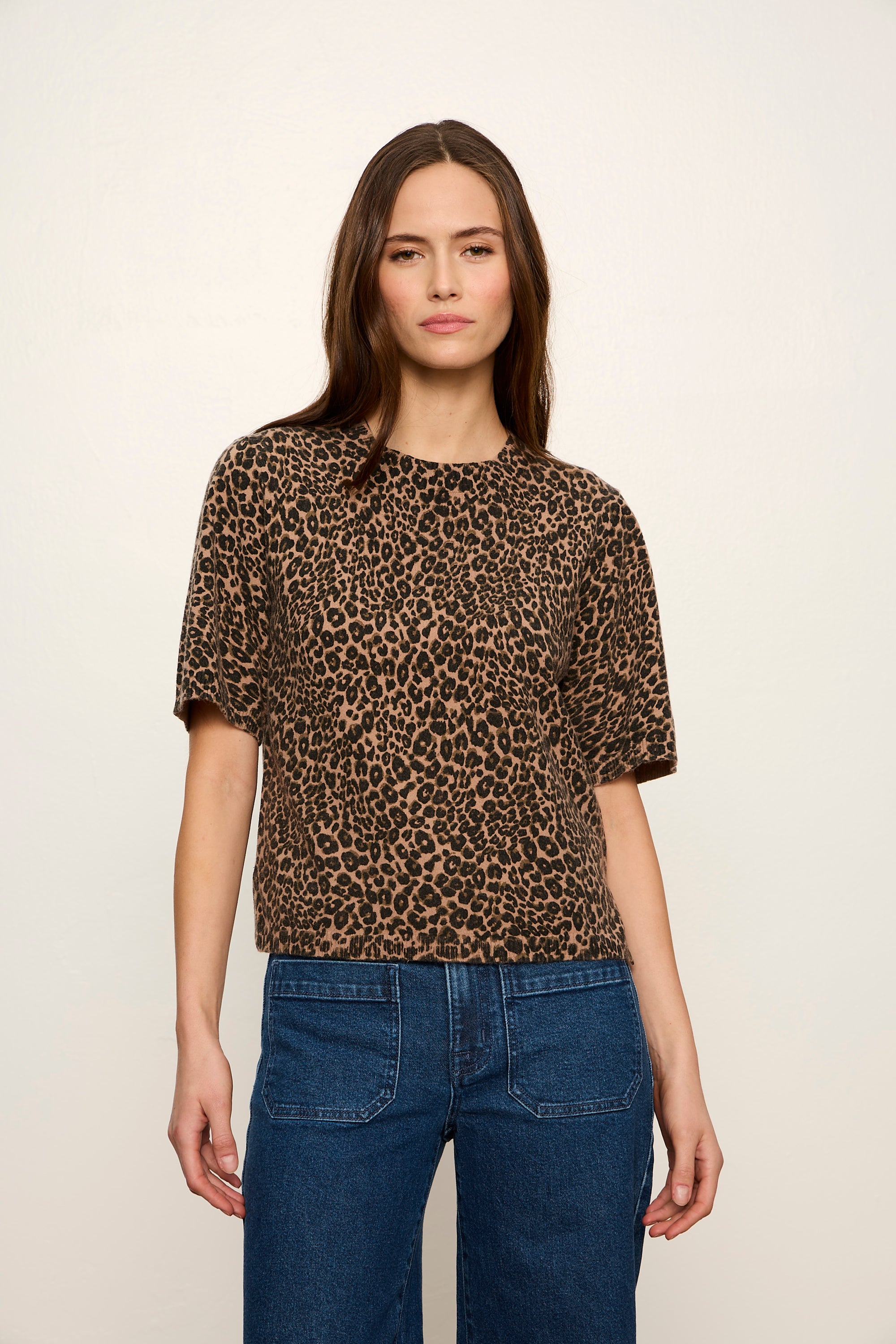 A woman modeling a short-sleeve leopard print top in brown and beige tones, paired with blue jeans, against a plain off-white studio background.