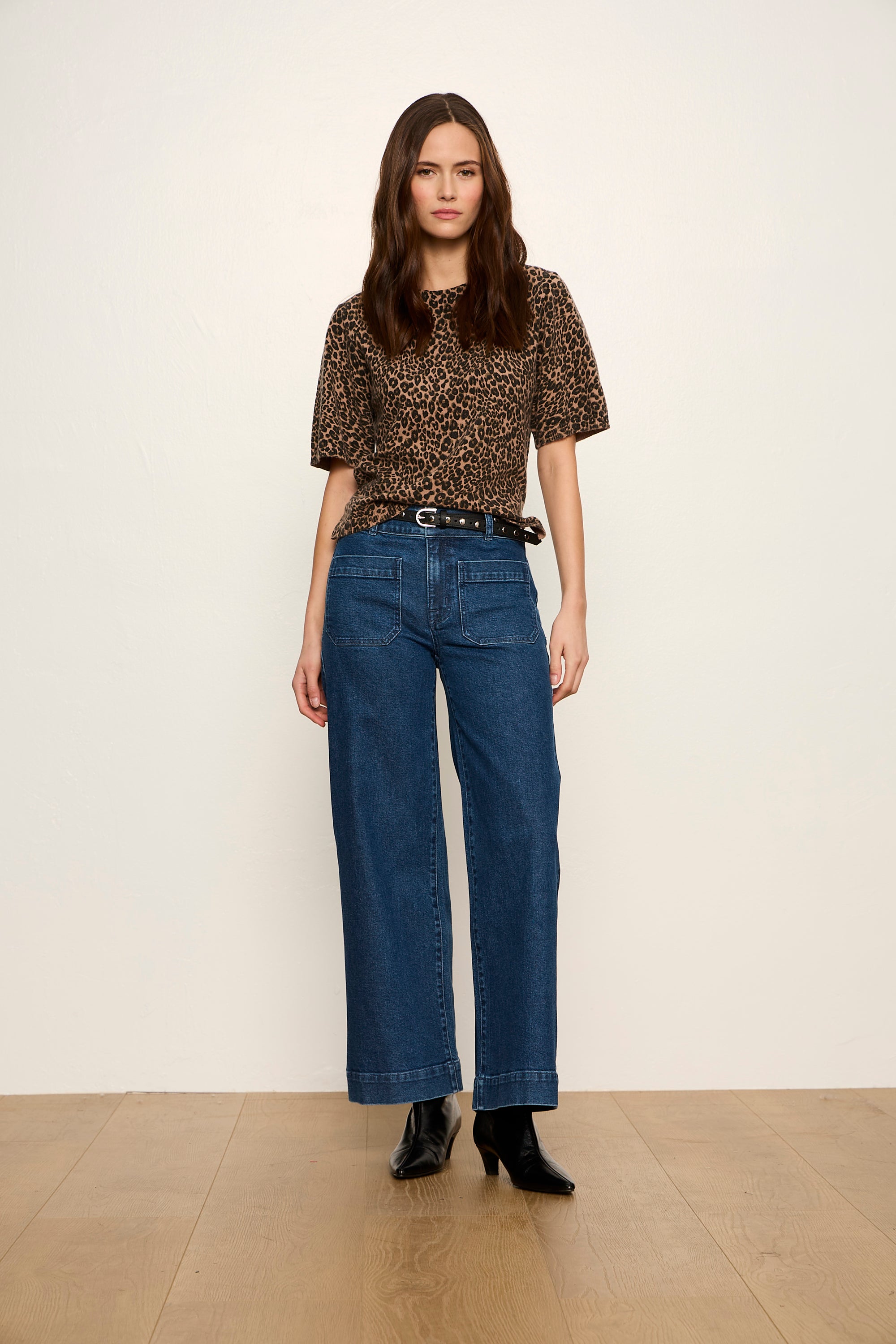 A woman modeling a short-sleeve brown and black leopard print top paired with high-waisted wide-leg blue jeans and black ankle boots, standing against a plain off-white wall and light wood floor.