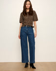A woman modeling a short-sleeve brown and black leopard print top paired with high-waisted wide-leg blue jeans and black ankle boots, standing against a plain off-white wall and light wood floor.
