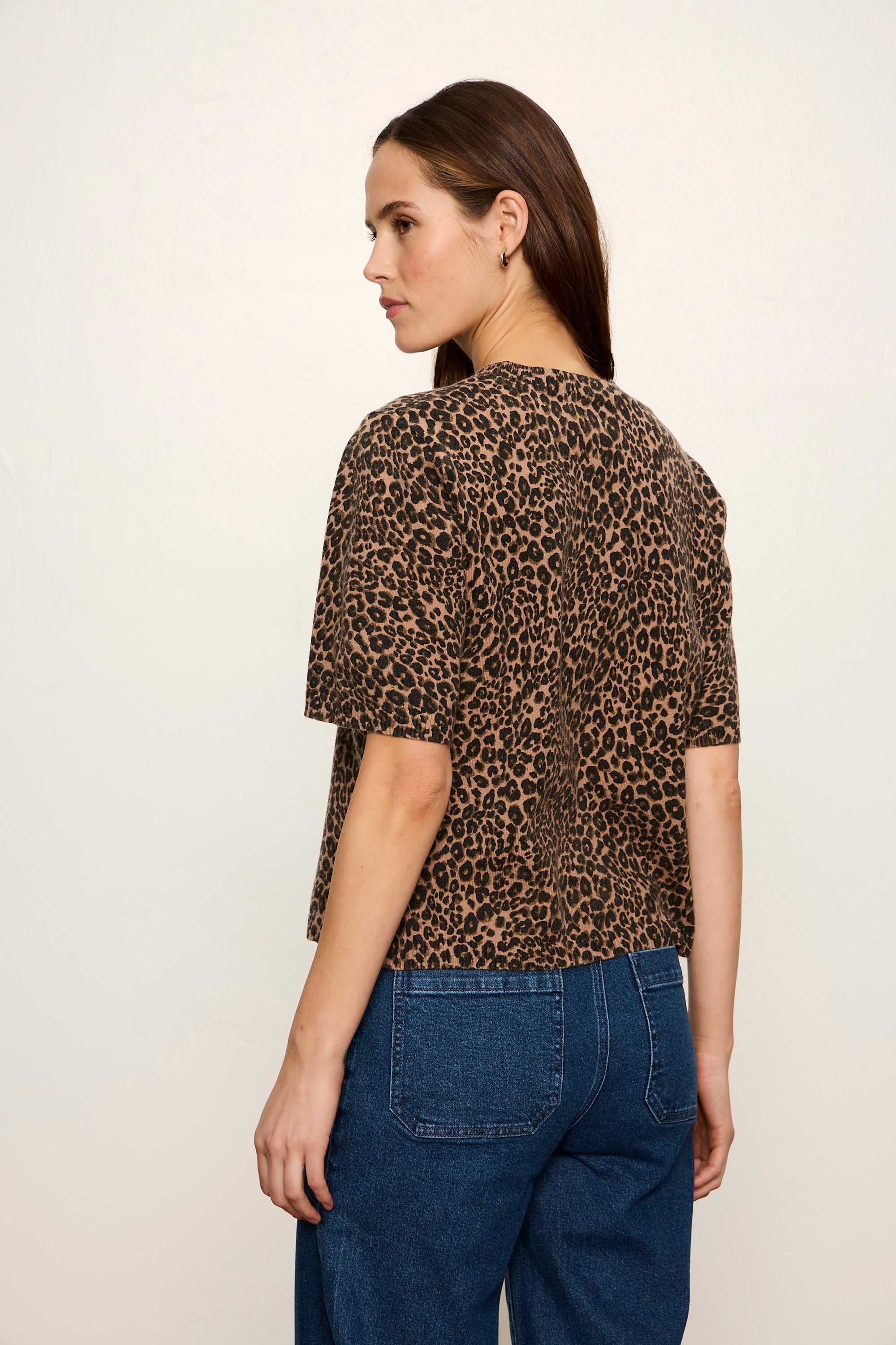A woman shown from the back wearing a brown and black leopard print short-sleeve top with blue jeans, photographed against a plain off-white studio background.