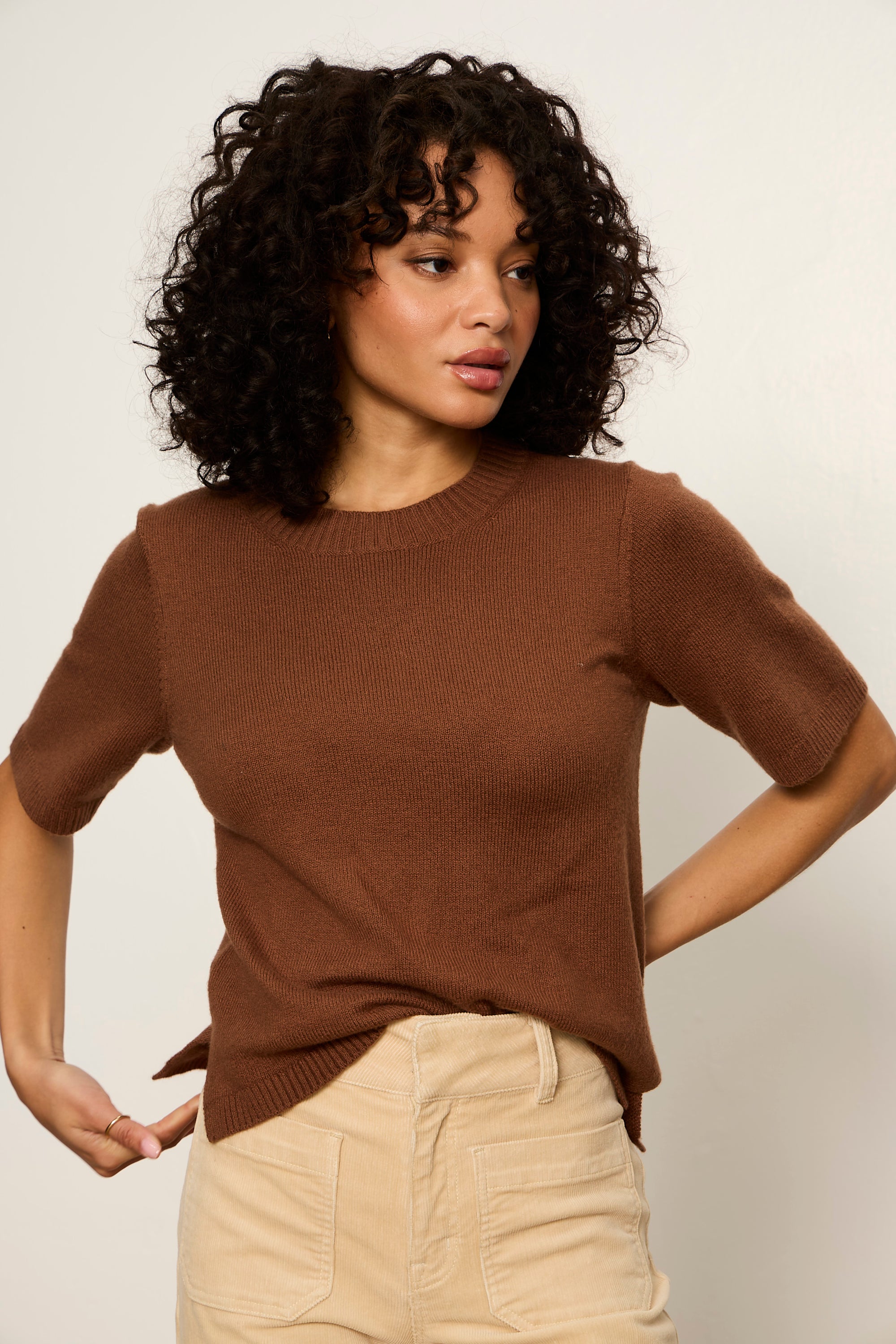 Woman wearing a brown sweater and beige pants against a plain background